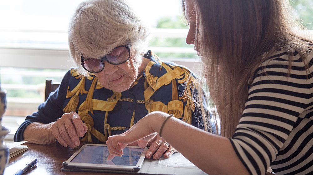 Two women sat together, one assisting the other with navigating an iPad.