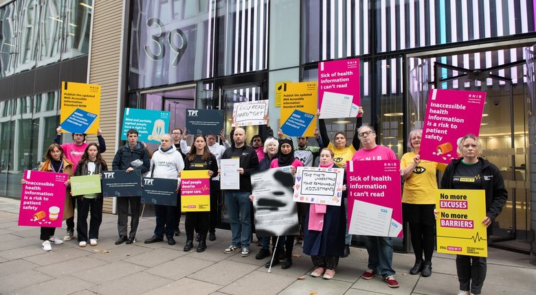 Photo of eighteen people stood outside of a glass fronted building. They are all holding a variety of placards in different colours, a mixture of yellow, pinks, and blues. Everyone has a serious expression.