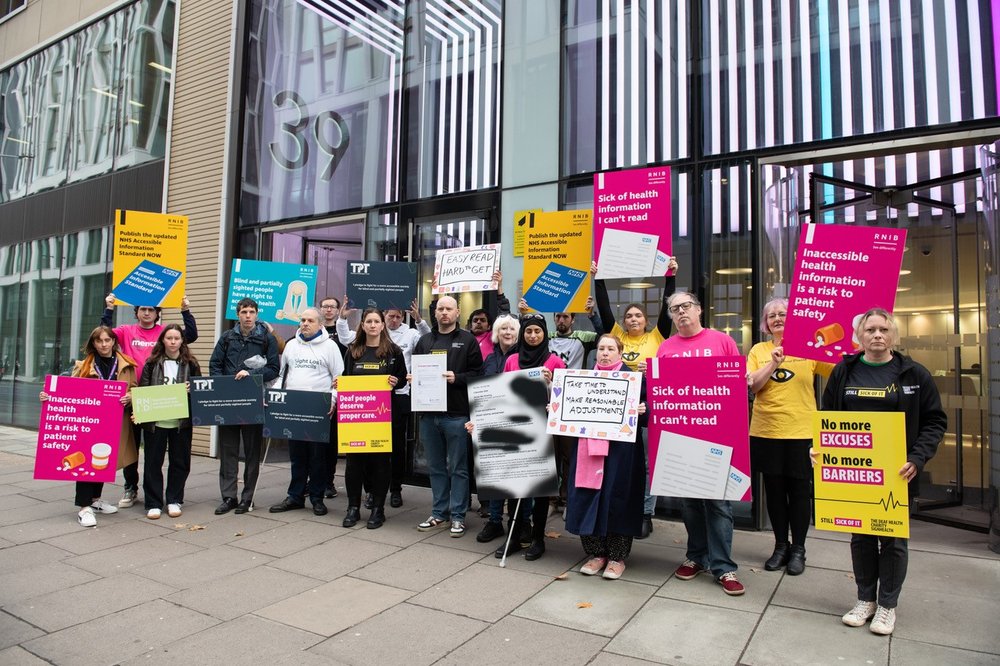 Photo of eighteen people stood outside of a glass fronted building. They are all holding a variety of placards in different colours, a mixture of yellow, pinks, and blues. Everyone has a serious expression.
