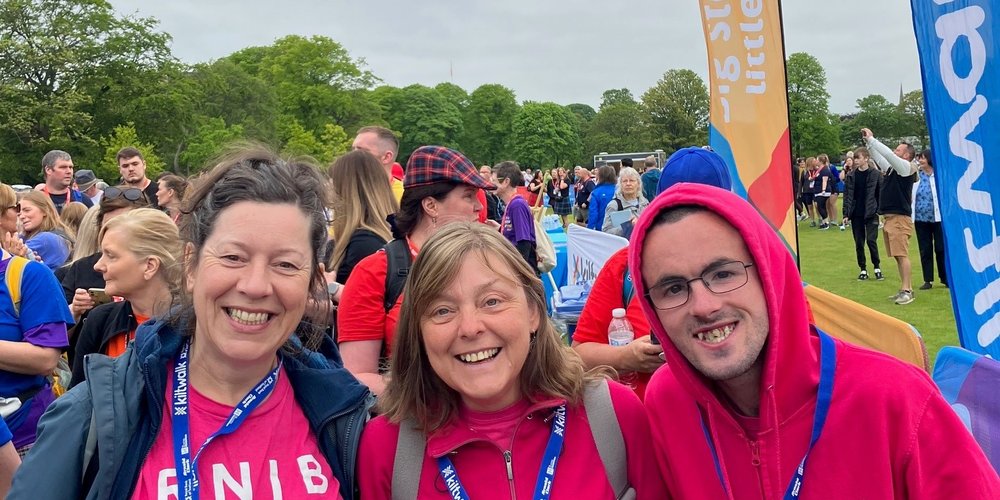Three people smiling at the camera wearing pink RNIB tops. They are outdoors completing a charity walk in Scotland. There are more people in the background but they aren't in focus.