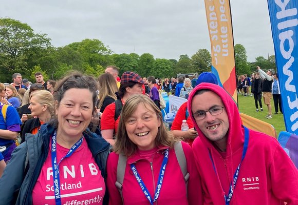 Three people smiling at the camera wearing pink RNIB tops. They are outdoors completing a charity walk in Scotland. There are more people in the background but they aren't in focus.