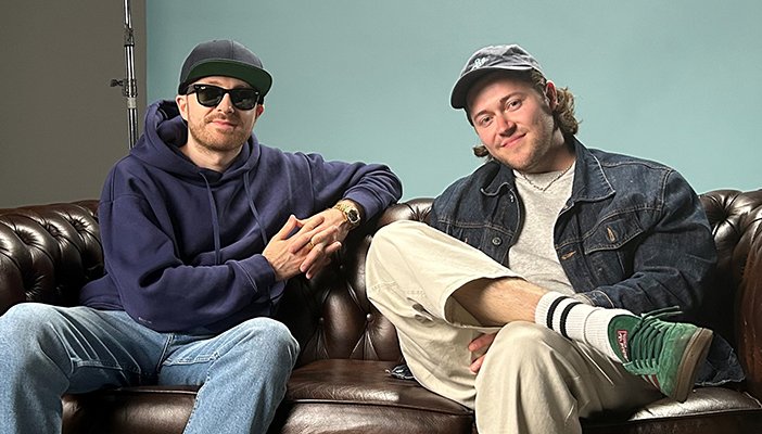 Two men sat on a sofa smiling at the camera