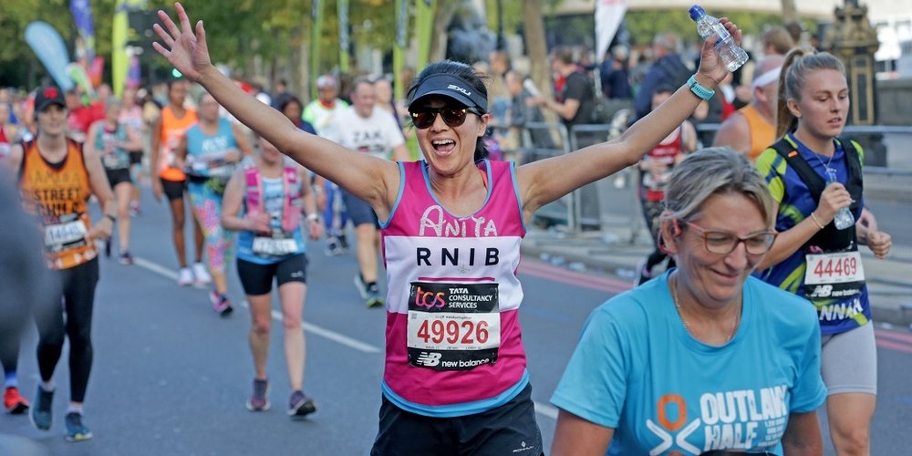 In the centre of the image Anita, a woman wearing a pink RNIB running vest with a number pinned to it, black shorts, a visor and sunglasses, is running the Hackney Half Marathon.  Her arms are wide and she has a big grin on her face as she looks towards the camera. She is surrounded by other people running the Hackney Half Marathon but they are not in focus.