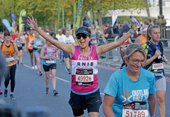 In the centre of the image Anita, a woman wearing a pink RNIB running vest with a number pinned to it, black shorts, a visor and sunglasses, is running the Hackney Half Marathon.  Her arms are wide and she has a big grin on her face as she looks towards the camera. She is surrounded by other people running the Hackney Half Marathon but they are not in focus.