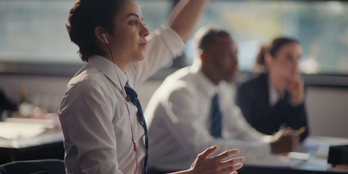 Girl sat down in a school classroom, dressed in her school uniform with her hand up.