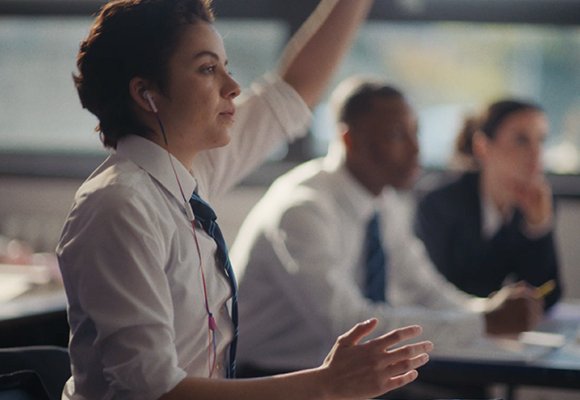 Girl sat down in a school classroom, dressed in her school uniform with her hand up.
