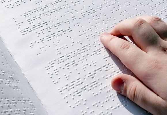 A closeup of a person’s hand as they read a braille book. Their hand is tracing along a sentence in the middle of the page.