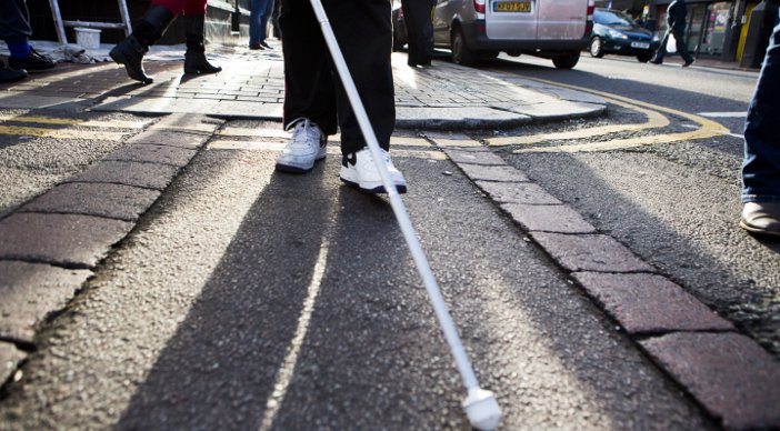 A person, from the knees down, crossing the road and using a white cane.