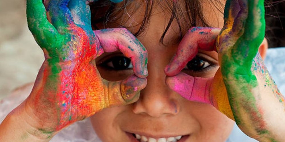 A child covered in multicoloured paint holding their hands up to their eyes like glasses and smiling.