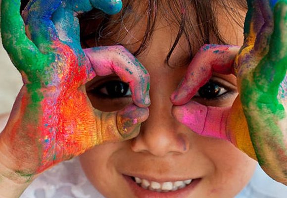 A child covered in multicoloured paint holding their hands up to their eyes like glasses and smiling.