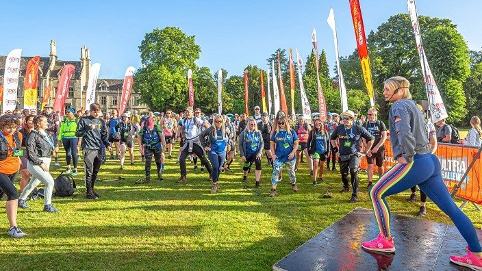 Image of a fitness instructor leading the warm up at the start of the Cotswold Way Challenge, surrounded by Cotswold stone buildings.