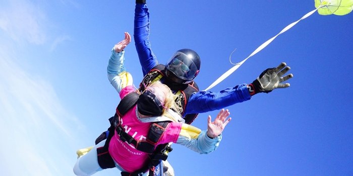 Two people taking part in a skydive, the one in front is wearing RNIB t-shirt, the one behind them is wearing a black helmet. Behind them is a bright blue sky.