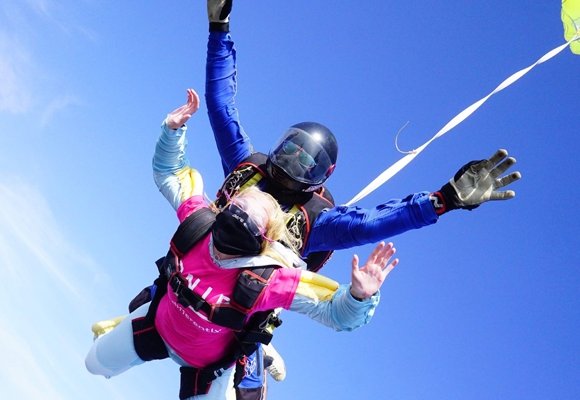 Two people taking part in a skydive, the one in front is wearing RNIB t-shirt, the one behind them is wearing a black helmet. Behind them is a bright blue sky.