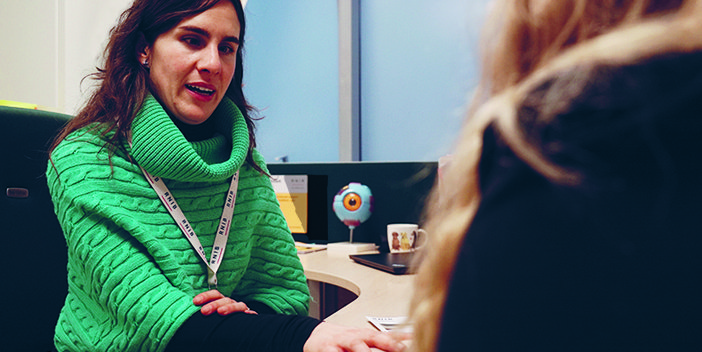 Eye Care Liaison Officer (ECLO) Alba, speaking to and holding the hand of a patient, who is out of focus to the side.