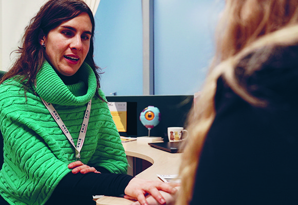 Eye Care Liaison Officer (ECLO) Alba, speaking to and holding the hand of a patient, who is out of focus to the side.