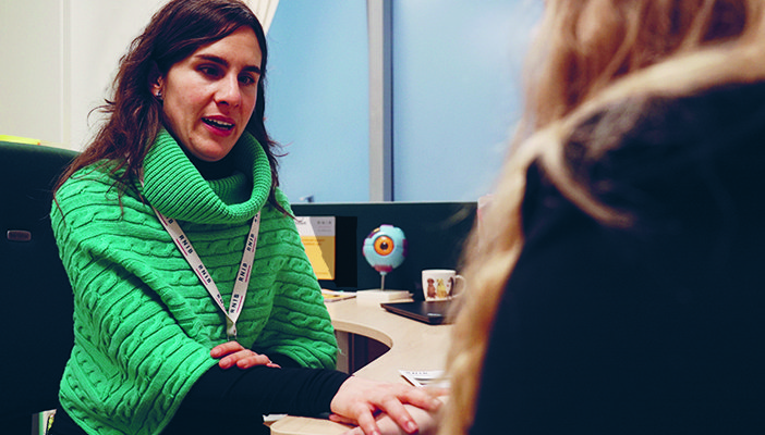 Eye Care Liaison Officer (ECLO) Alba, speaking to and holding the hand of a patient, who is out of focus to the side.
