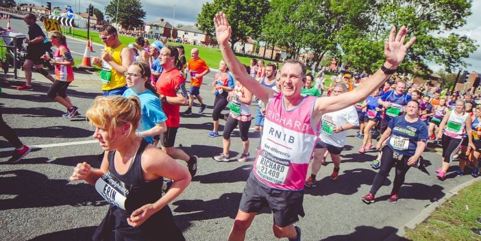 An image of lots of runners taking part in the Great North Run. At the centre of the image one older man holds his arms out wide and smiles at the camera. He is wearing a pink RNIB running vest and black shorts.