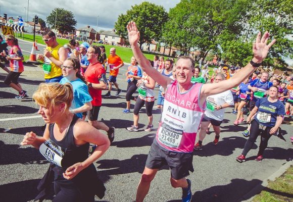An image of lots of runners taking part in the Great North Run. At the centre of the image one older man holds his arms out wide and smiles at the camera. He is wearing a pink RNIB running vest and black shorts.