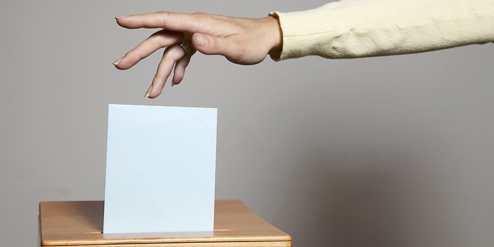 A hand dropping a folded ballot into a ballot box.