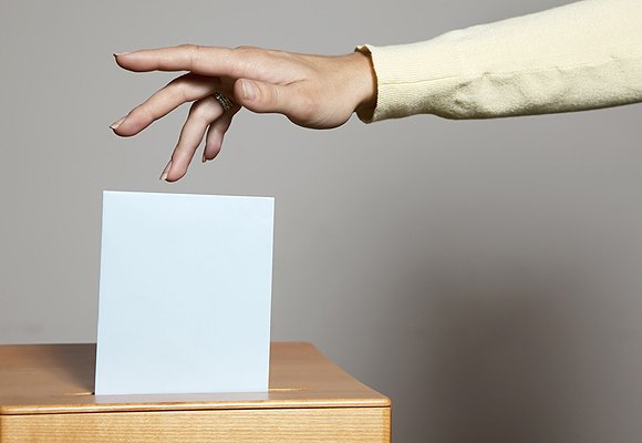 A hand dropping a folded ballot into a ballot box.