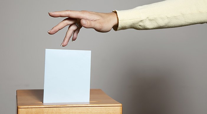 A hand dropping a folded ballot into a ballot box.
