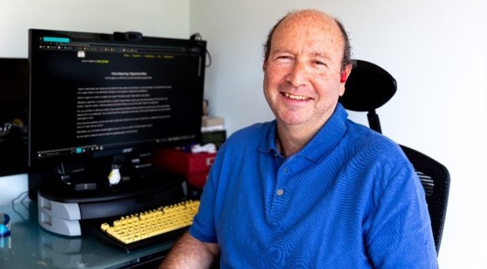 Jonathan sits in a desk chair wearing a blue polo shirt and smiling, his desk and computer are just behind him and the screen shows a website in inverted colours