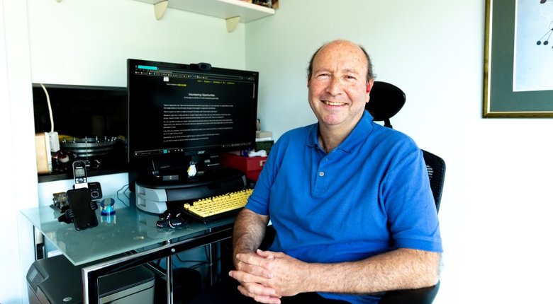 A man sits at his desk with his chair turned towards the camera smiling, he is using dark mode on his computer screen.