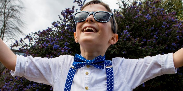A young boy in dotty sunglasses and clothes, smiling, with his arms outstretched towards the sky.