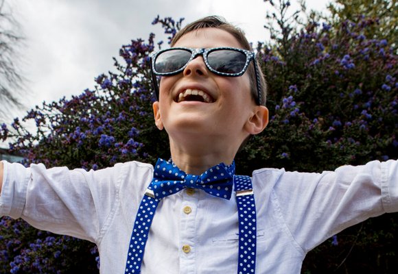 A young boy in dotty sunglasses and clothes, smiling, with his arms outstretched towards the sky.