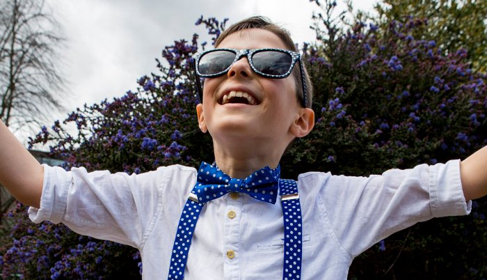 A young boy in dotty sunglasses and clothes, smiling, with his arms outstretched towards the sky.