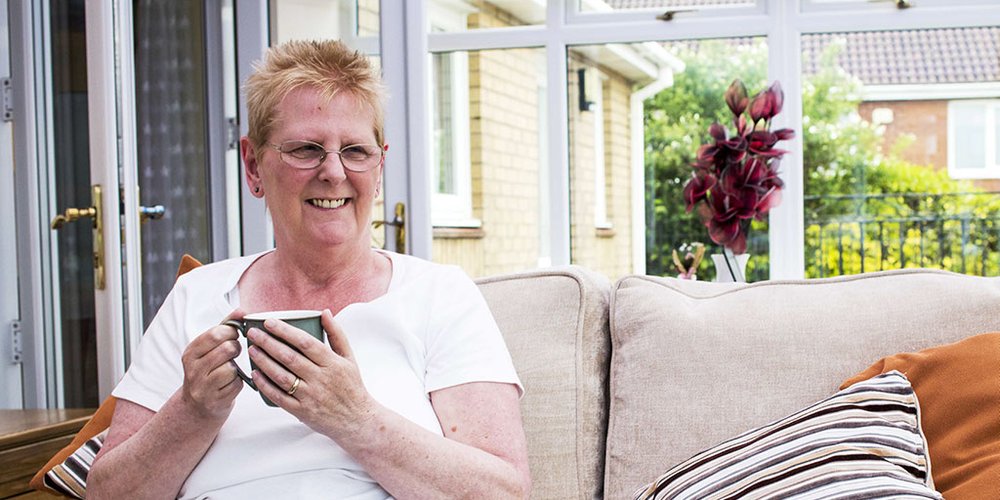 Lady sat on a sofa in a conservatory enjoying a cup of tea.