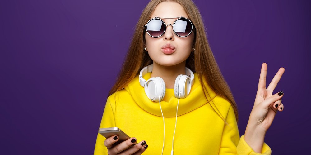 Lady in bright yellow dress wearing round sunglasses and white headphones round her neck connected to her phone in one hand. She is making a peace sign with the other hand.