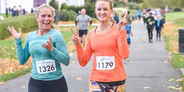 Two ladies running at the Leeds Running Festival event. One is wearing a turquoise coloured top and the other is wearing a bright orange coloured top and is giving the peace sign with her hands.