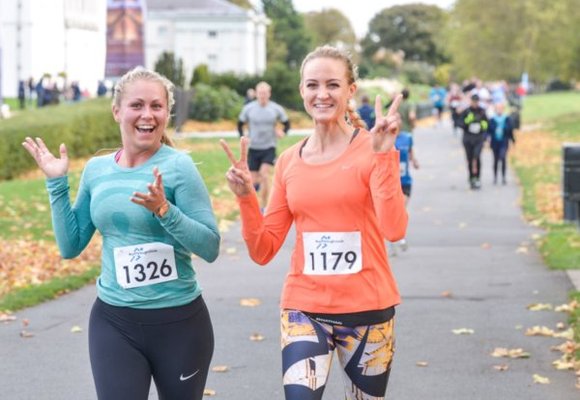 Two ladies running at the Leeds Running Festival event. One is wearing a turquoise coloured top and the other is wearing a bright orange coloured top and is giving the peace sign with her hands.