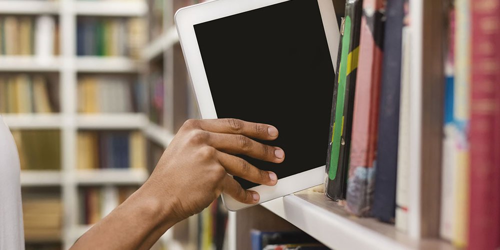 A person taking a tablet off a shelf filled with books.