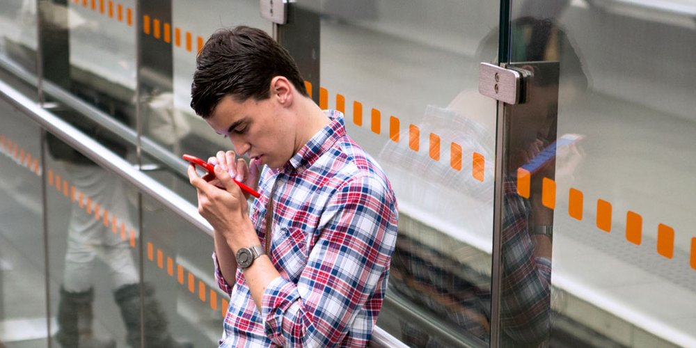 A young man standing at a bus stop reading information on a red smartphone.