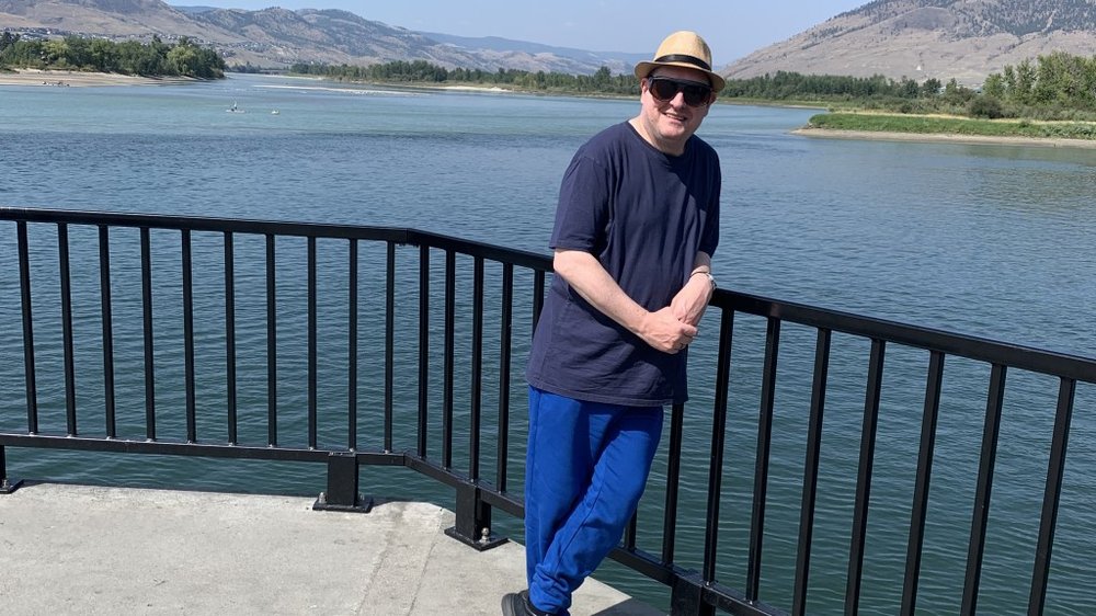 A person leaning against some railings smiling at the camera with a lake in the background on sunny day.