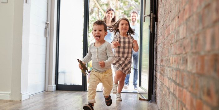 A young boy and girl running happily through the open front door of a house while their parents look on behind them from outside.