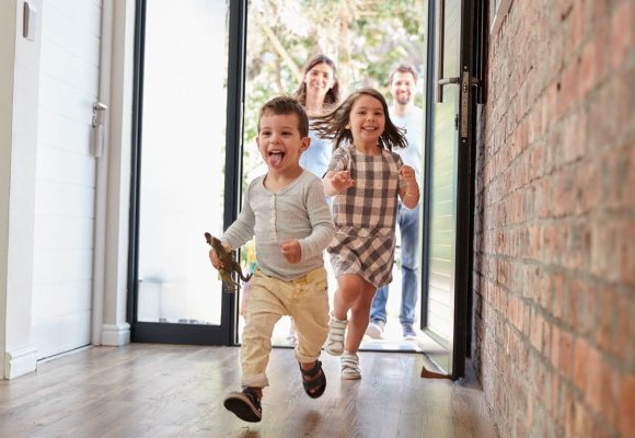A young boy and girl running happily through the open front door of a house while their parents look on behind them from outside.