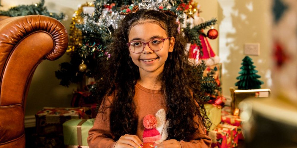 Remi holding a wrapped present, sat in front of a Christmas tree covered in tinsel and baubles.