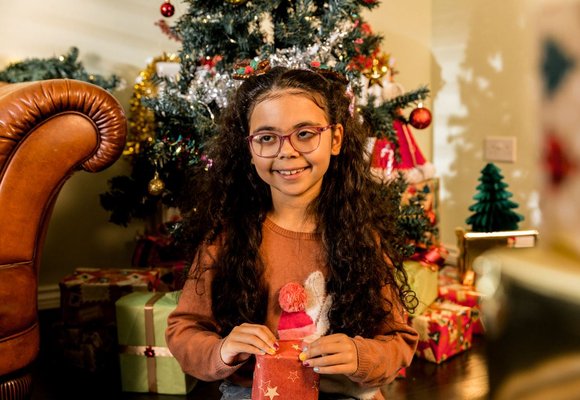 Remi holding a wrapped present, sat in front of a Christmas tree covered in tinsel and baubles.