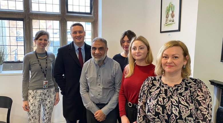 Eleanor and Maqsood in grey tops, Wes Streeting in a dark suit and overcoat, Gen in a red top and Vivienne in a black and white patterned dress standing in a group in the corner of a bright coffee shop looking happy