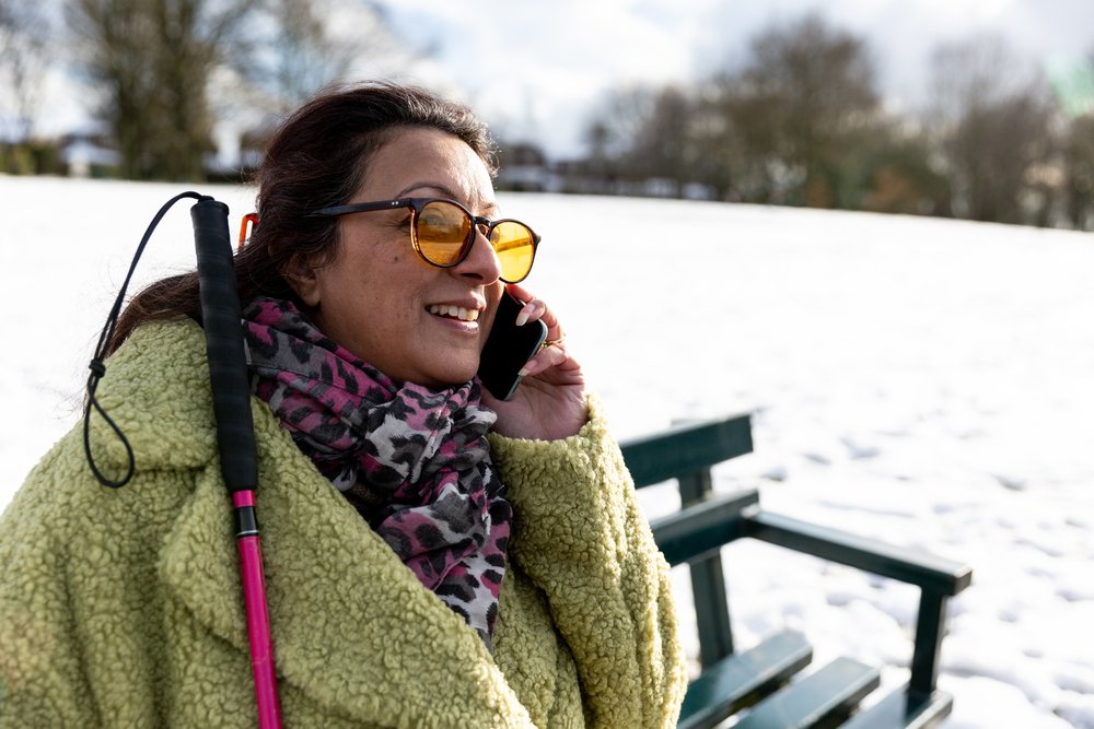 Perm sat outside on bench in the snow, holding walking cane, smiling while on the phone.
