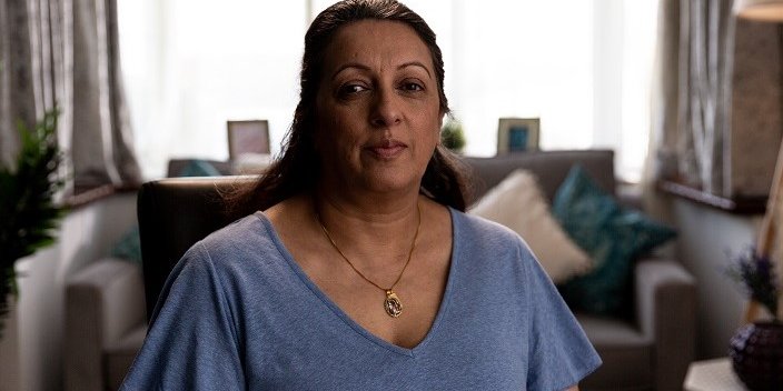 Image of lady sat in the foreground lounge with a sofa, window and lampshade in the background. She’s looking into the camera with a neutral expression.