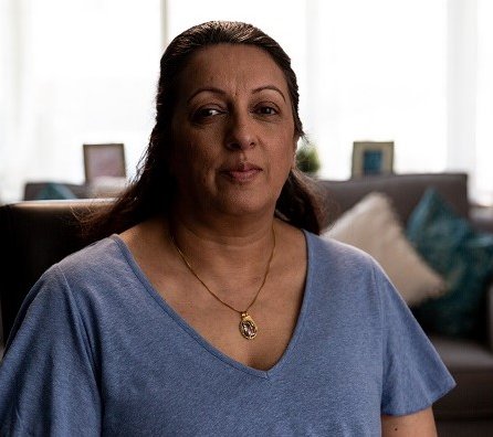 Image of lady sat in the foreground lounge with a sofa, window and lampshade in the background. She’s looking into the camera with a neutral expression.