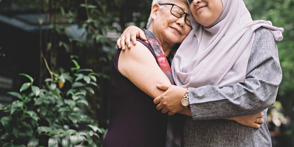 Two women in a caring embrace standing in front of lush vegetation