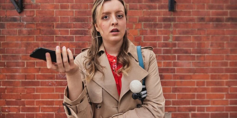 A woman with sight loss in a brown coat against a brick wall, holding her phone and a cane.