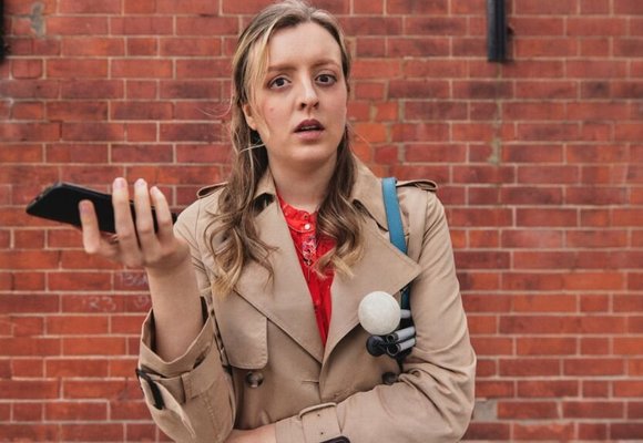 A woman with sight loss in a brown coat against a brick wall, holding her phone and a cane.