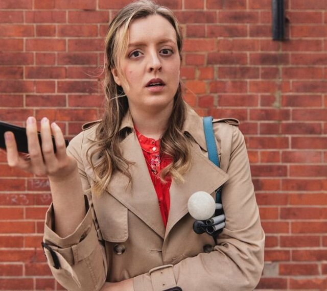 A woman with sight loss in a brown coat against a brick wall, holding her phone and a cane.