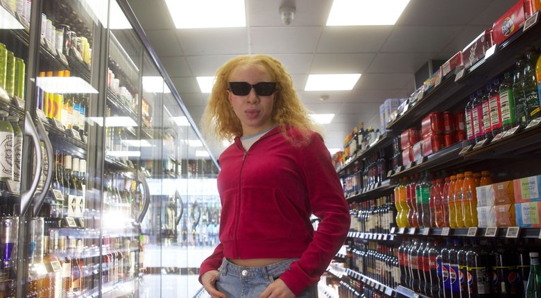 A sight impaired woman standing in the aisle of a shop wearing sunglasses and a trendy pink hoodie and jeans